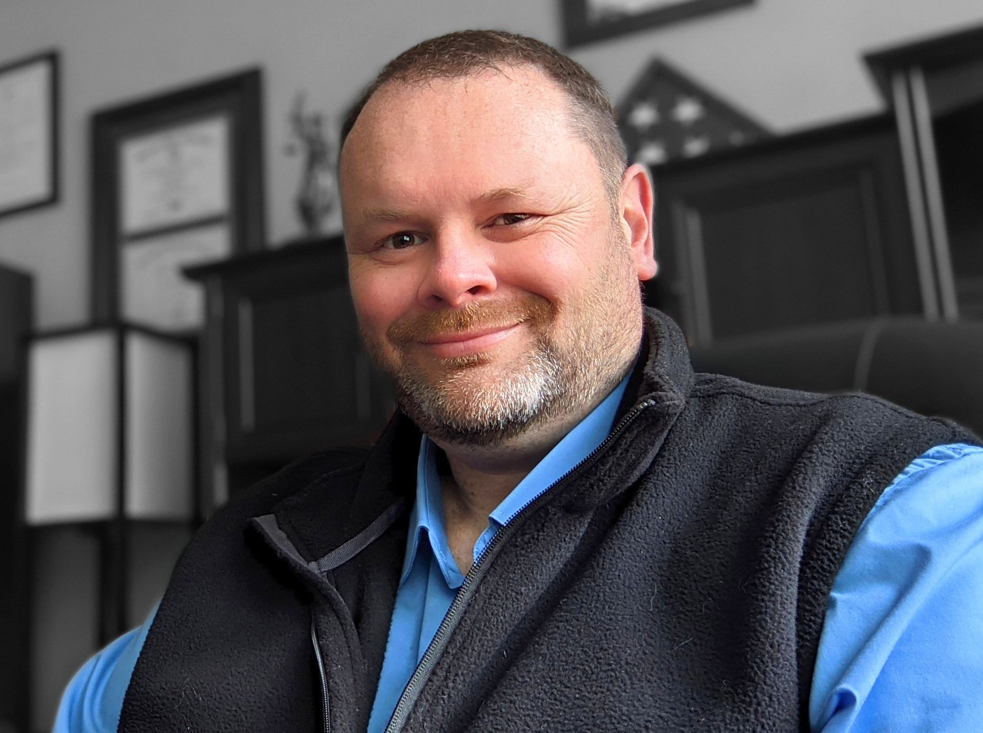 A man wearing a blue shirt and a black vest is smiling for the camera.