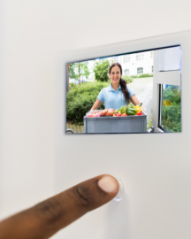 A close-up of a finger pressing a smart doorbell display showing a person holding a crate of fresh produce at the door.
