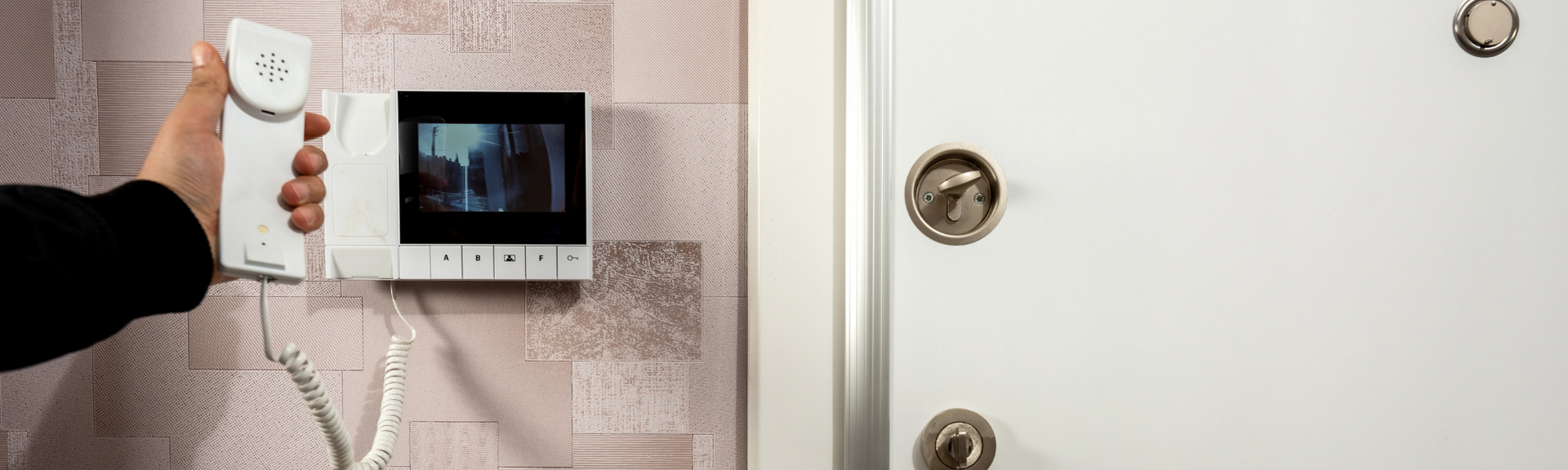 A hand holding a white intercom handset near a wall-mounted video monitor next to a white door with locks.