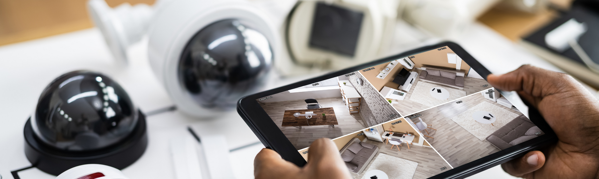 A person holds a tablet displaying a live multi-camera security feed next to several security camera units on a desk.