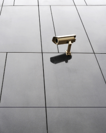 A security camera mounted on a modern wall with large grey panels and thin black grout lines.
