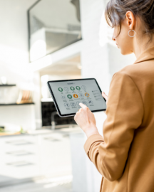 A person in a brown jacket uses a tablet to control a smart home system in a bright, modern kitchen.