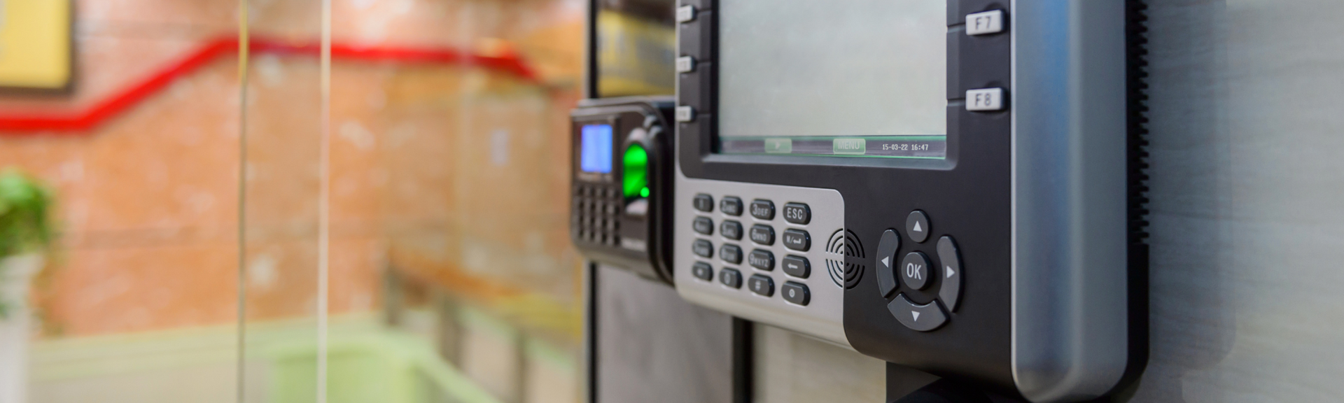 A biometric security terminal with a keypad and fingerprint scanner mounted on a wall in a blurred indoor office setting.