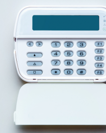 A white wall-mounted security alarm keypad with a digital display and numeric buttons, shown with its cover flipped down.