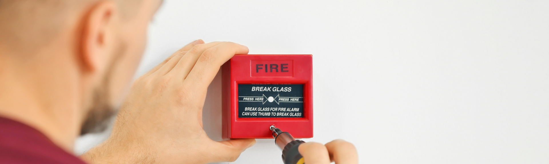 A person uses a screwdriver to install a red fire alarm pull station on a white wall.