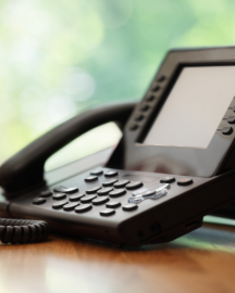 A black VoIP desk phone sits on a wooden surface against a soft-focus, bright green background.