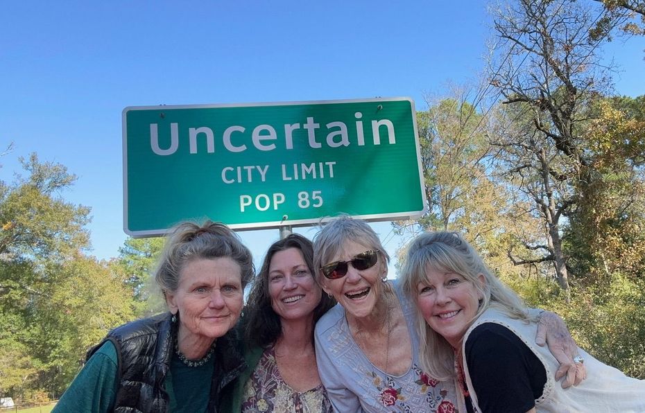 Four women are posing for a picture in front of a green sign that says uncertain city limit pop 85