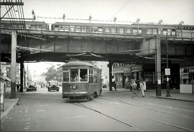 Vintage streetcar travels under an elevated train track. Pedestrians walk on a city street; buildings line the road.