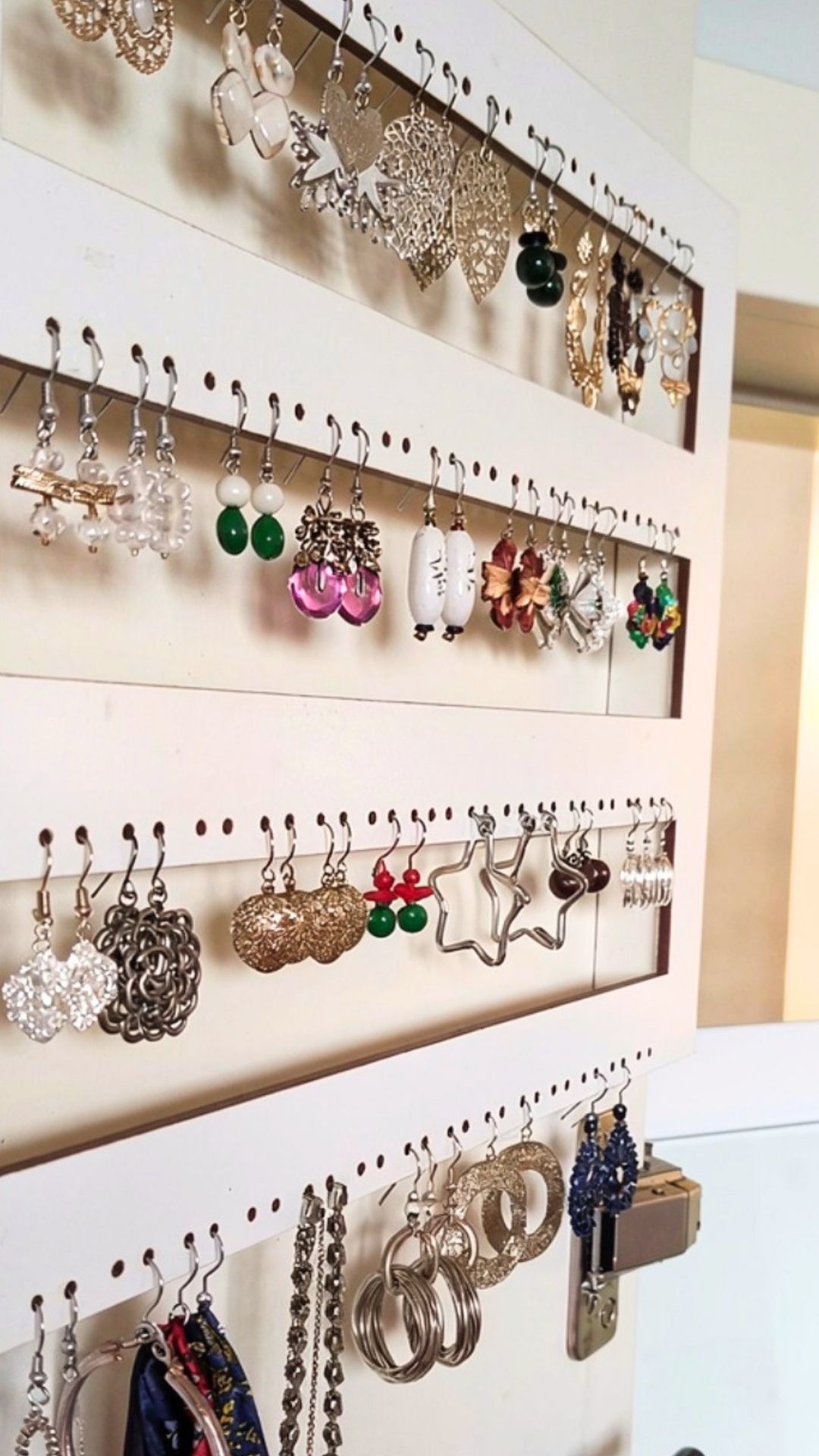 Exhibición de pendientes y joyas colgantes en estantes blancos perforados, con piezas variadas de plata, verde, rosa y oro.