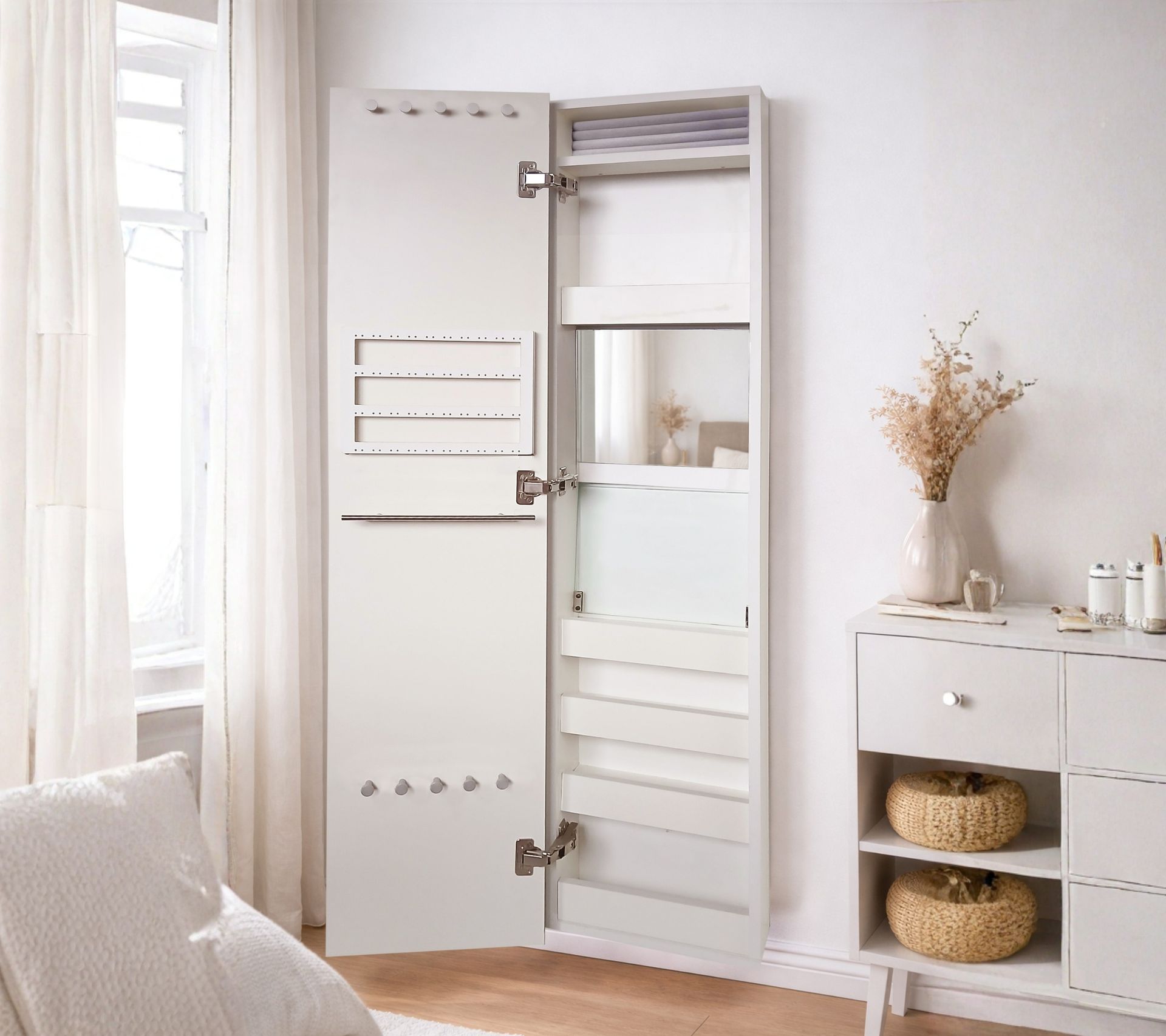 Armario blanco con puerta de espejo en una habitación luminosa, junto a una cortina y un aparador con cestas y flores secas.