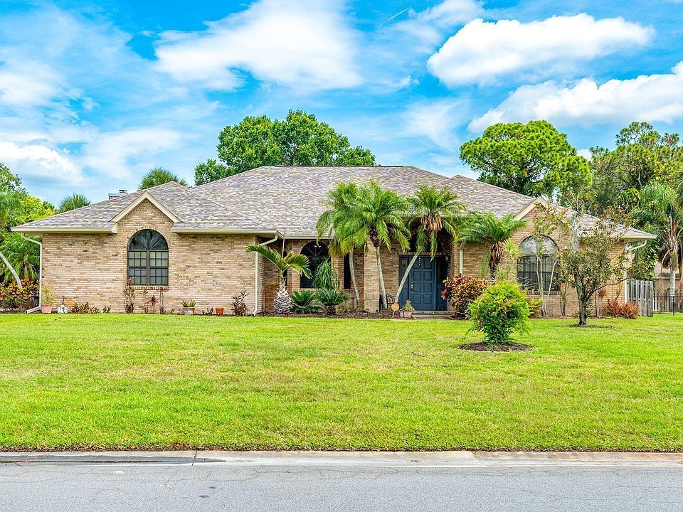 Single-story brick house with palm trees and lush green lawn under a blue sky.