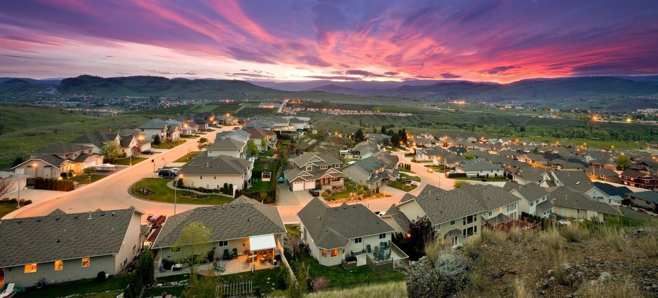 Aerial view of a residential neighborhood at sunset, vibrant pink and purple sky over houses, hills in background.