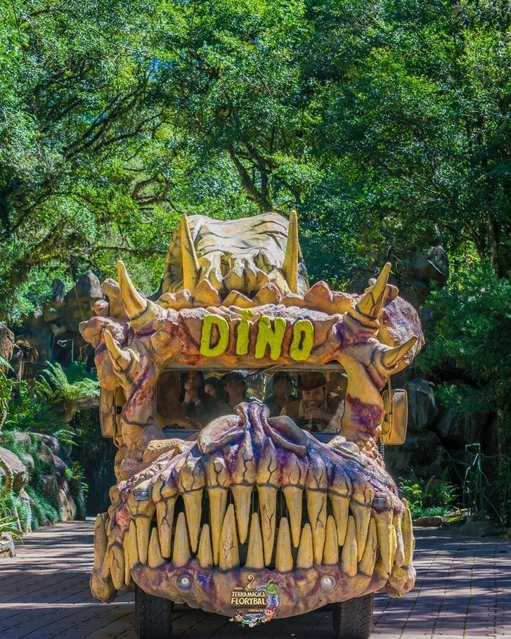 Um caminhão de dinossauro está estacionado em um parque com árvores ao fundo.