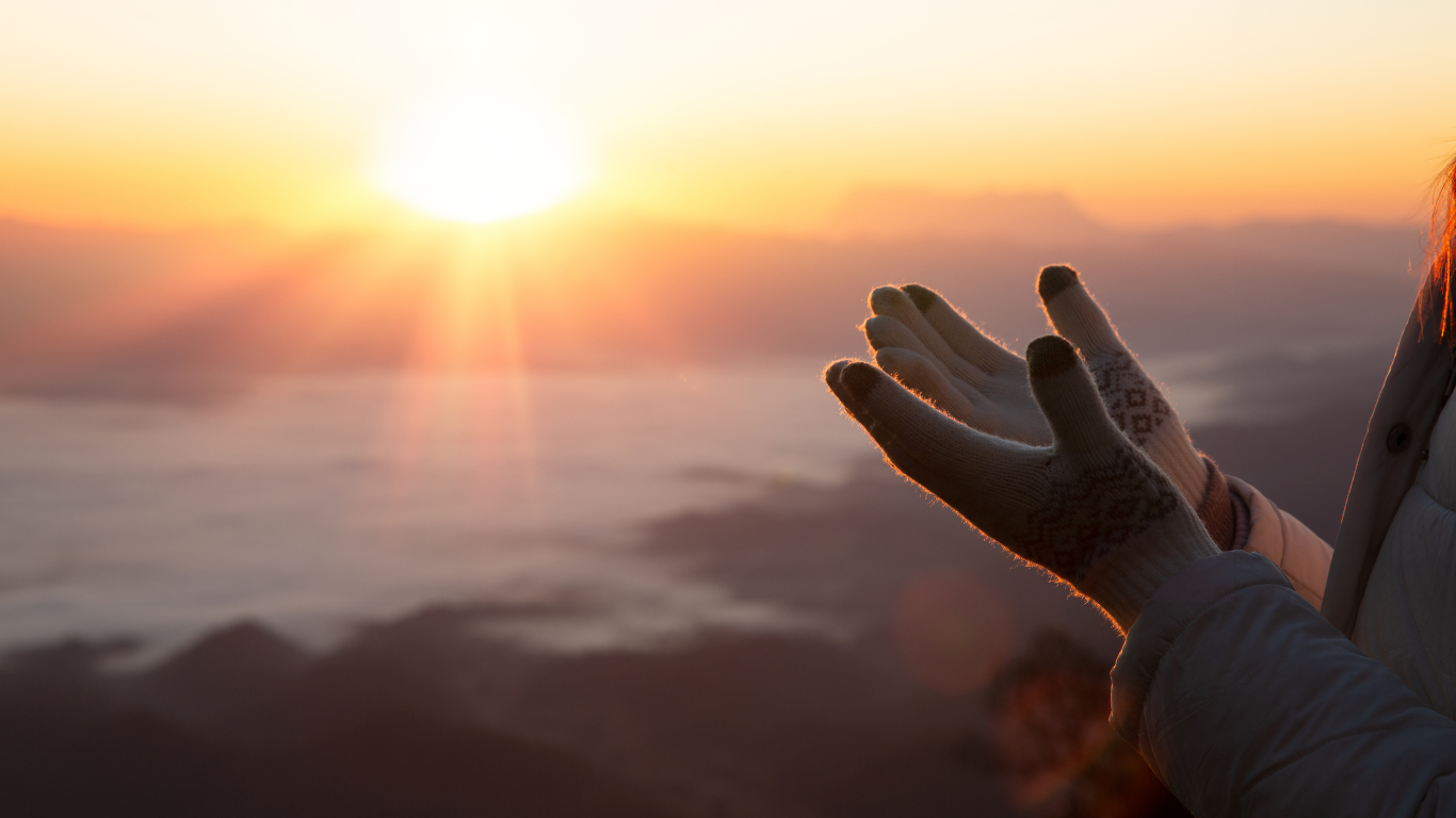 Hands outstretched toward a sunrise over a mountainous landscape. Warm light bathes the scene.