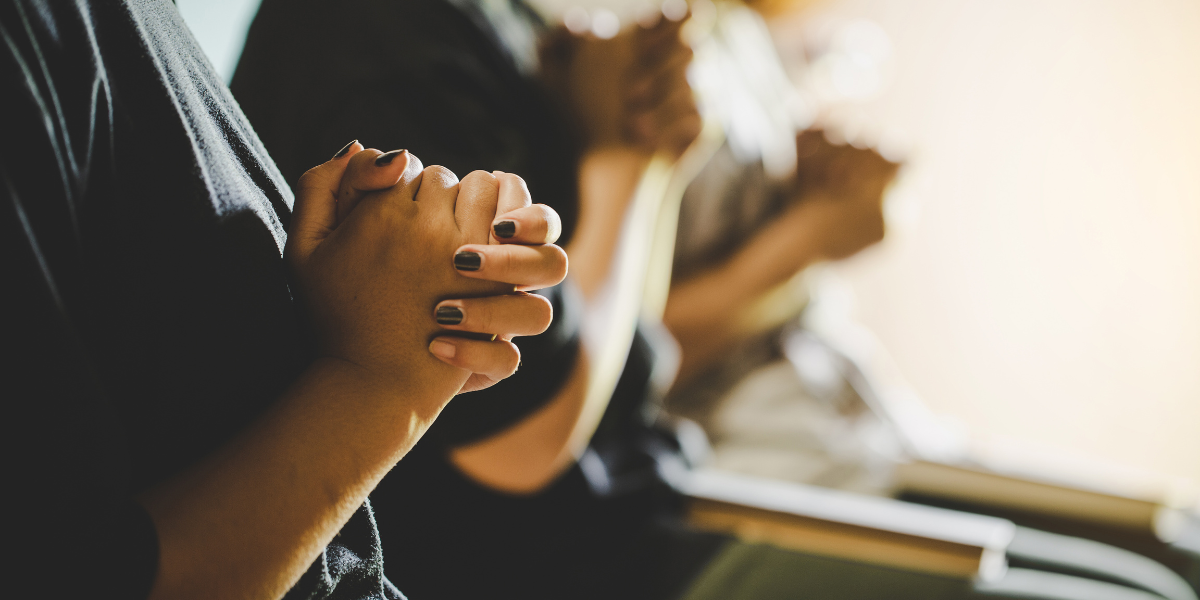 Hands clasped in prayer, in a group setting with soft lighting.