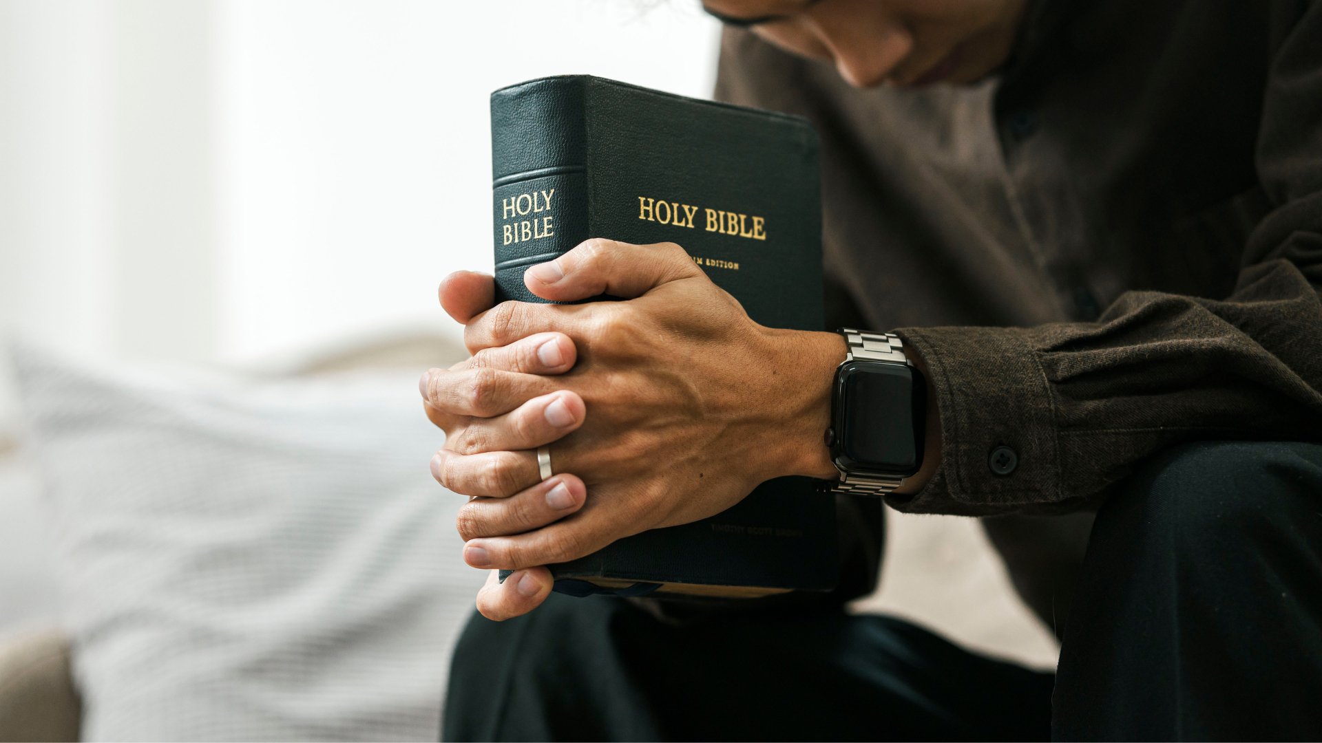 Person with hands clasped around a Bible, head bowed in prayer, near a window.