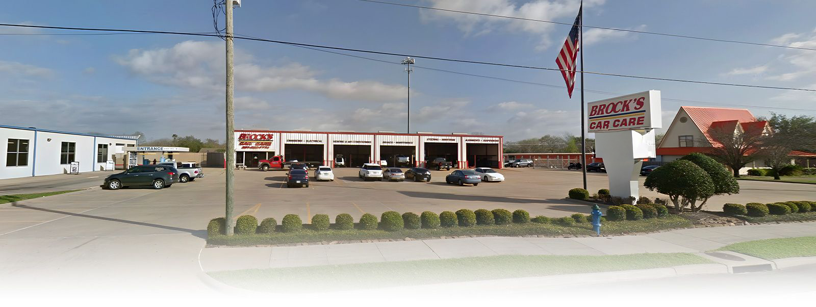 A commercial auto repair shop with multiple service bays, a parking lot, and a branded sign under an American flag.