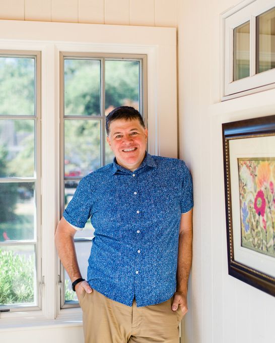 A man in a blue shirt is leaning against a wall next to a window.