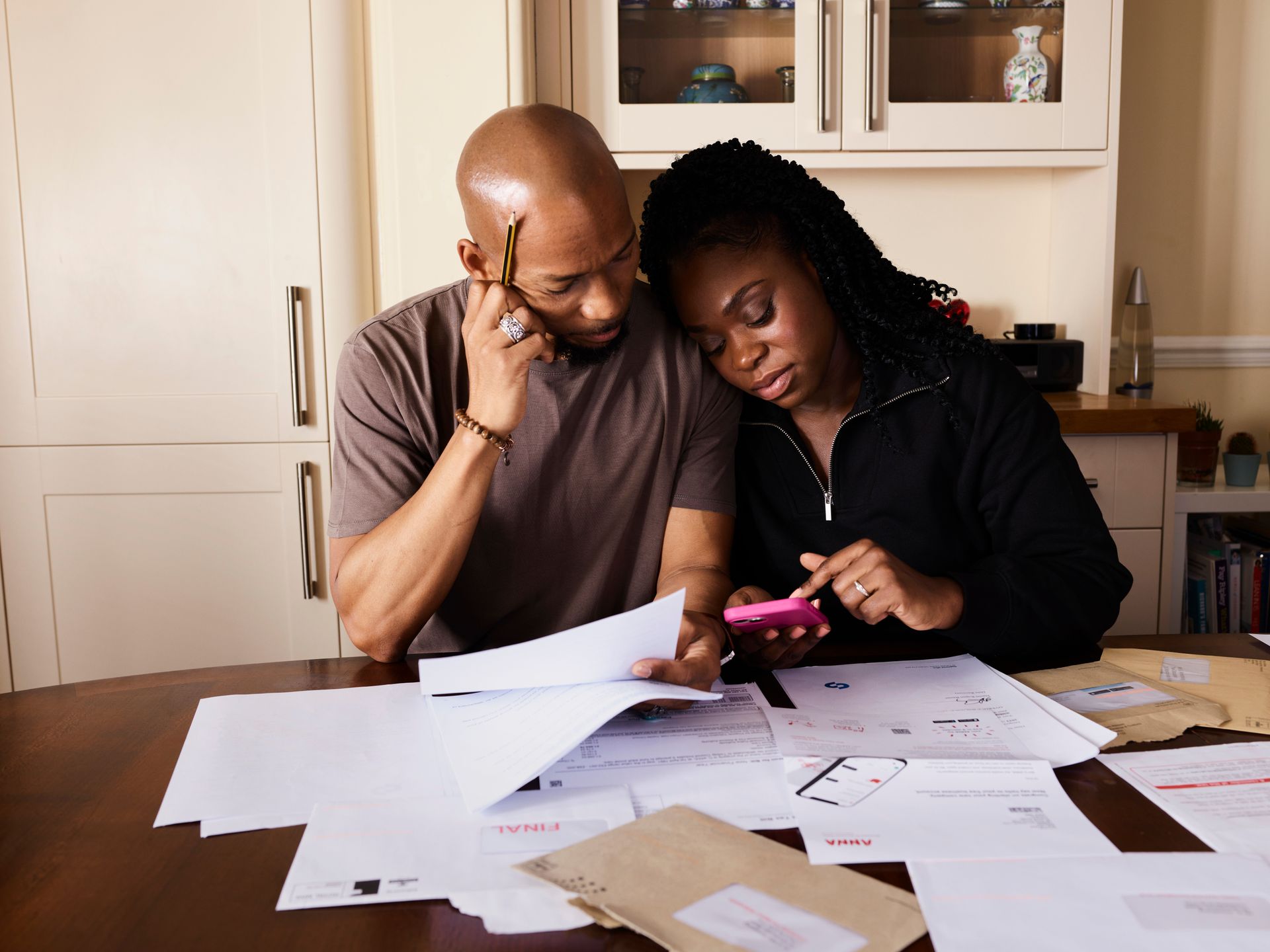 Two people reviewing papers at a kitchen table, one using a pink phone, with documents spread out.