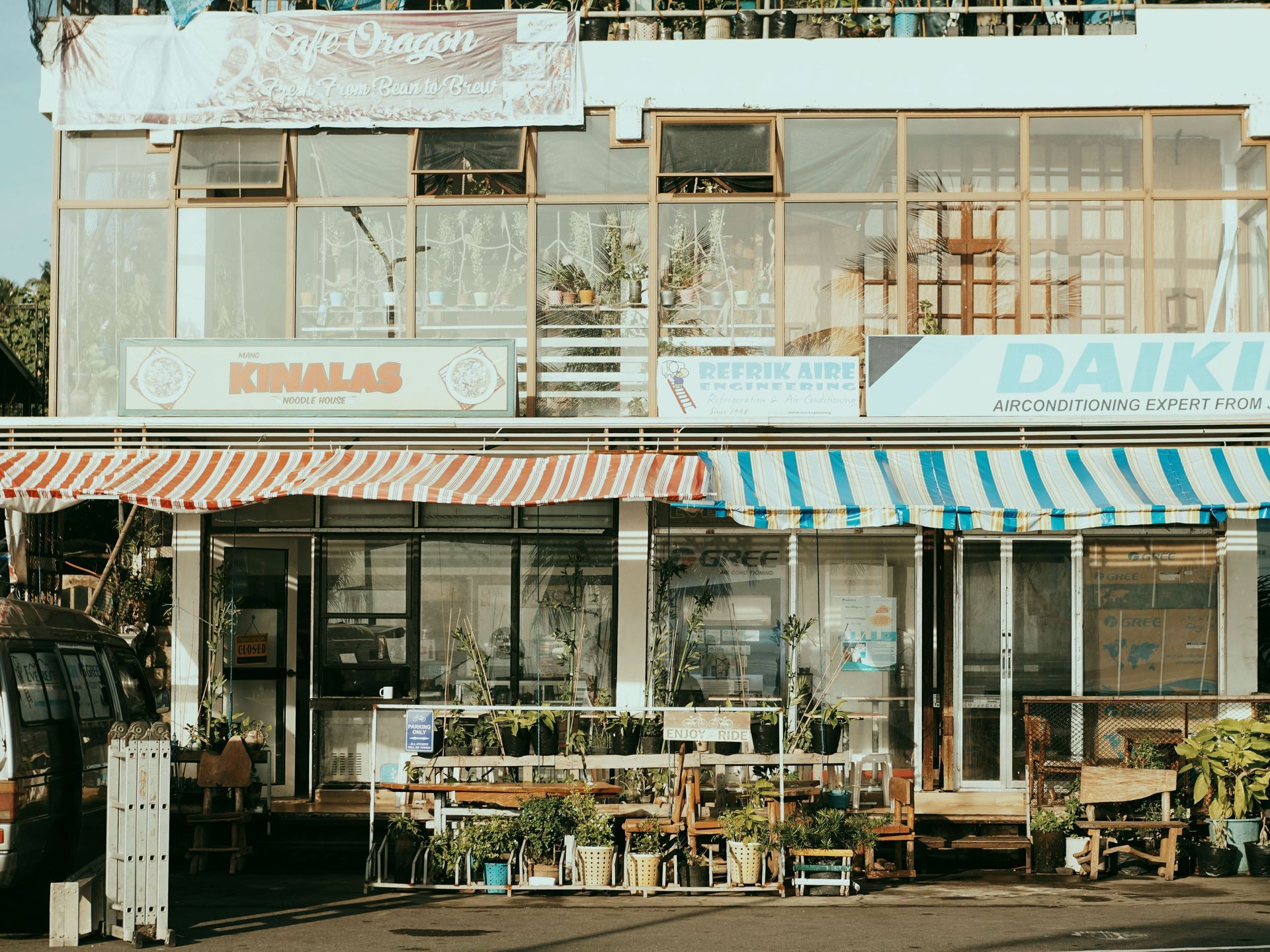 Small storefronts with striped awnings, potted plants, and a two-story building facade on a sunny street.