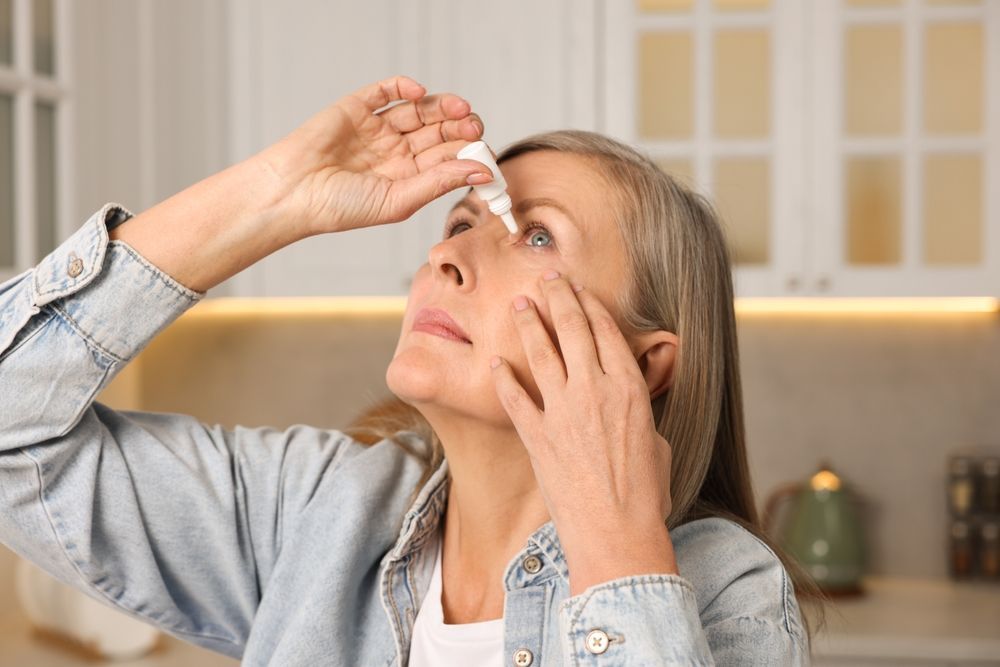 An elderly woman is applying eye drops to her eyes.