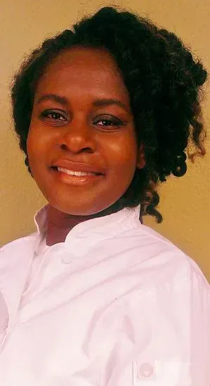 Woman in white chef's coat smiles, curly dark hair, neutral background.