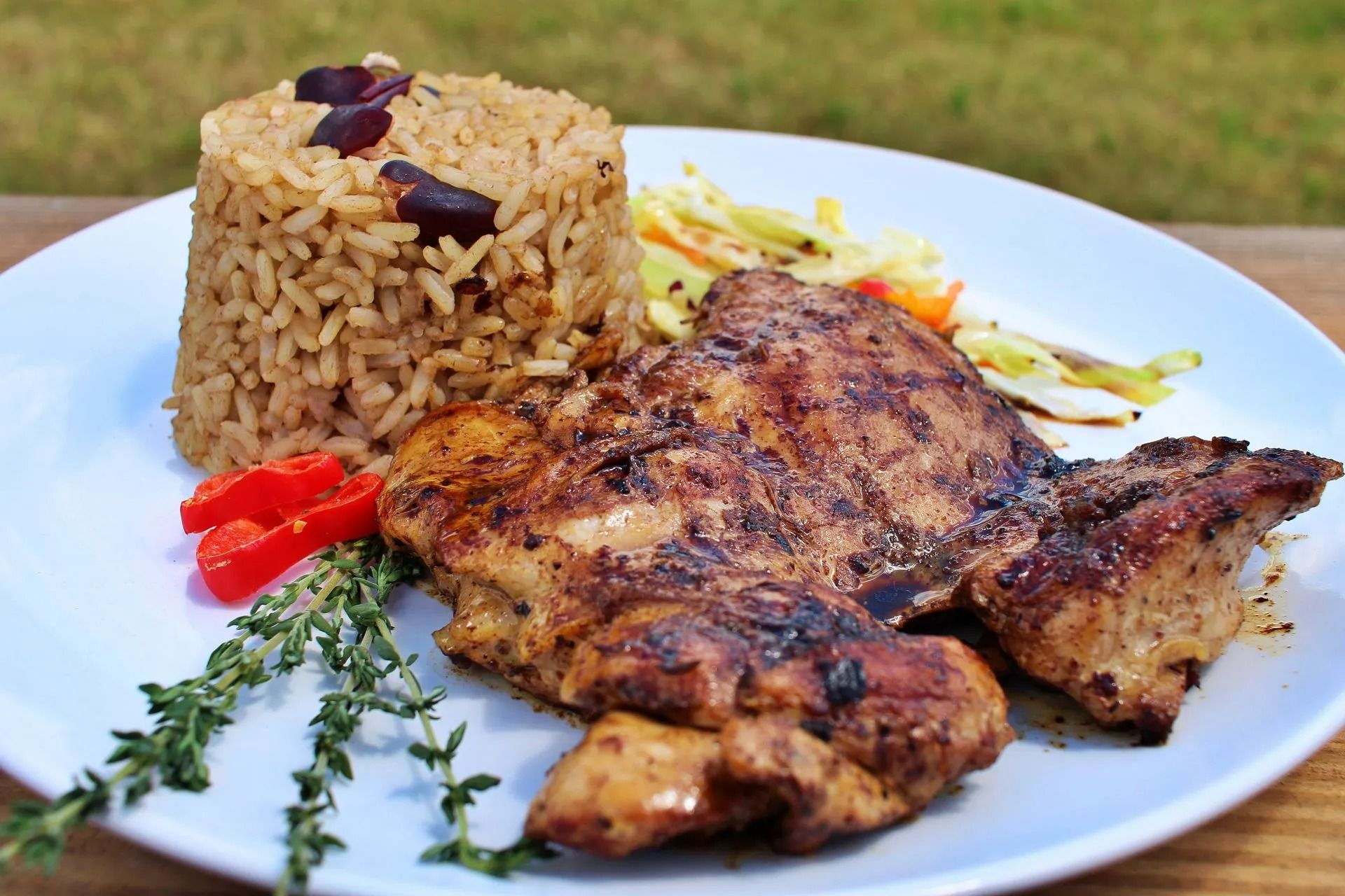 Grilled chicken, rice, and slaw on a white plate with fresh thyme and a red pepper, outdoor setting.