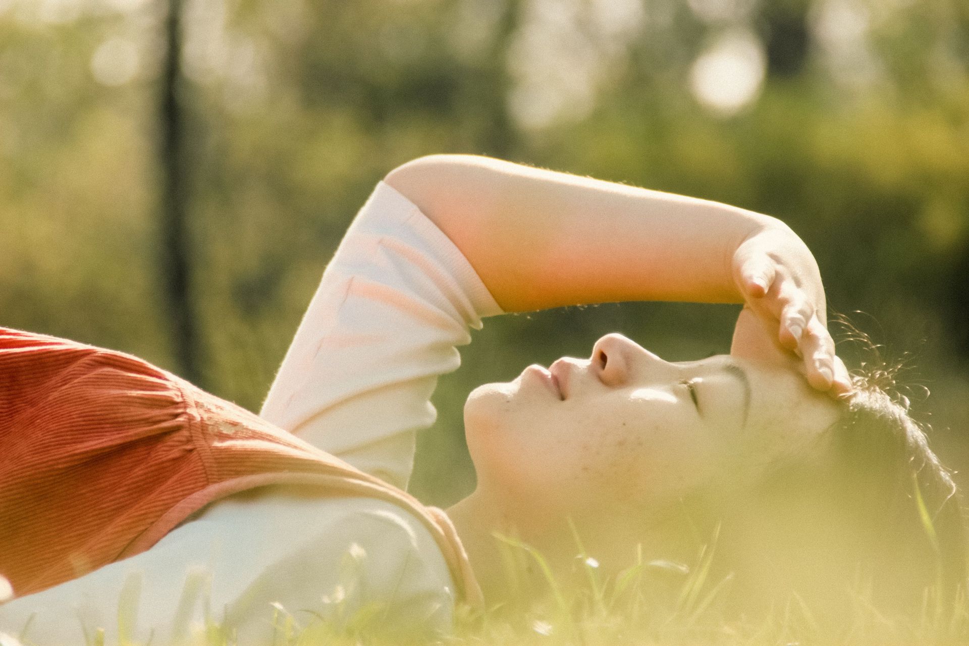 Person lying in sunlit grass, arm over face, with soft green background