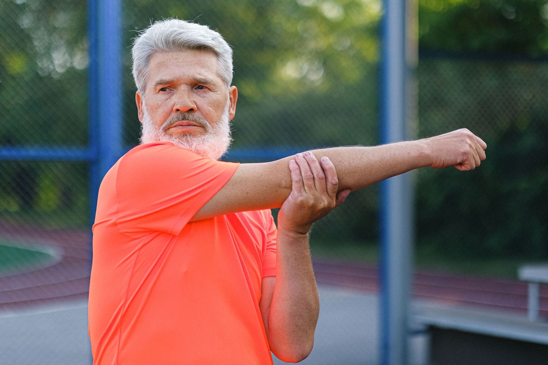 Man in bright orange shirt stretching his arm on an outdoor sports court