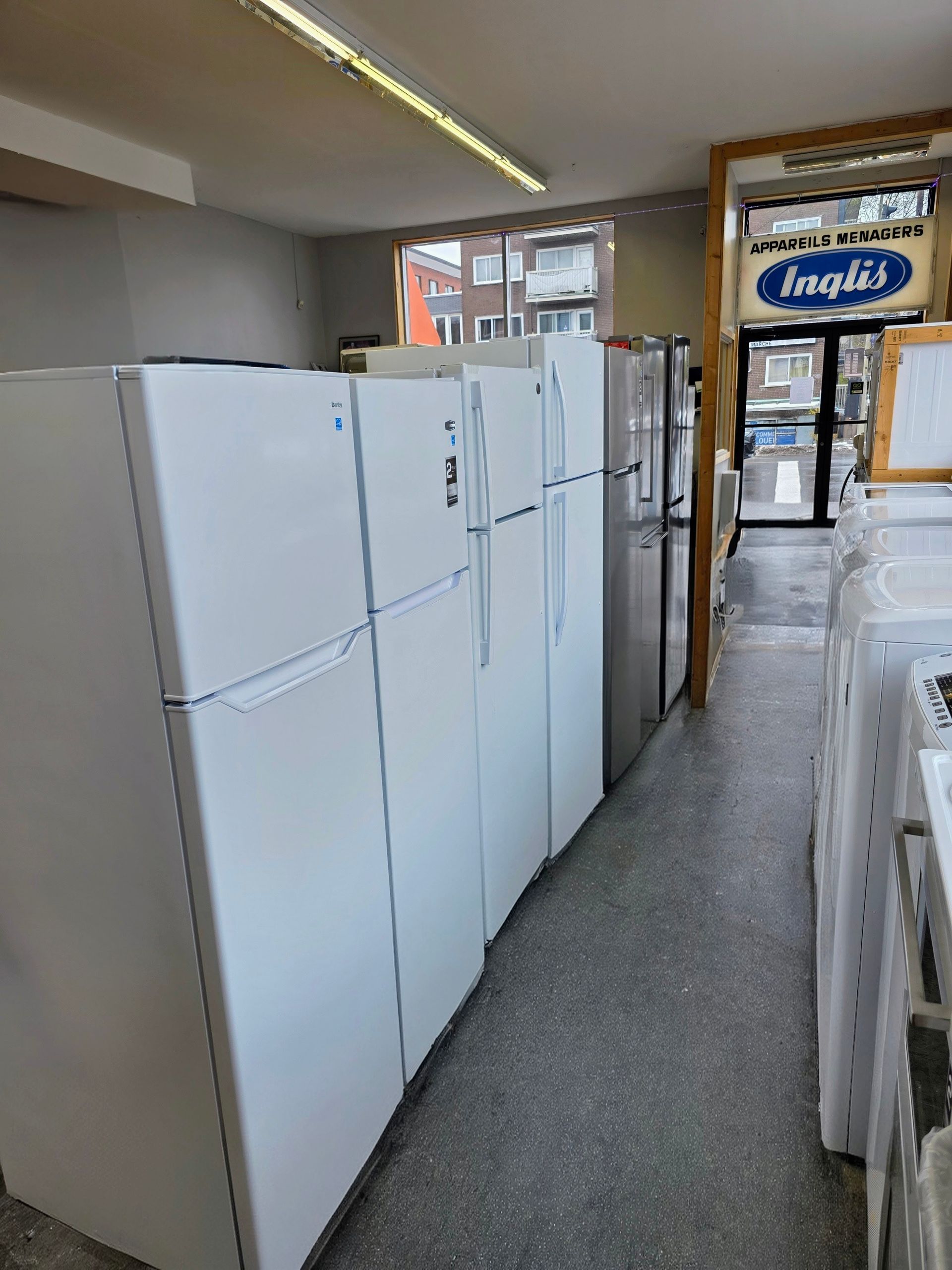 Des réfrigérateurs alignés dans un magasin d'électroménager, modèles blancs et argentés, vue sur les allées, éclairage fluorescent.