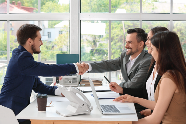 Man in a suit shakes hands with a man in a gray suit at a desk, with two other people looking on in an office setting.