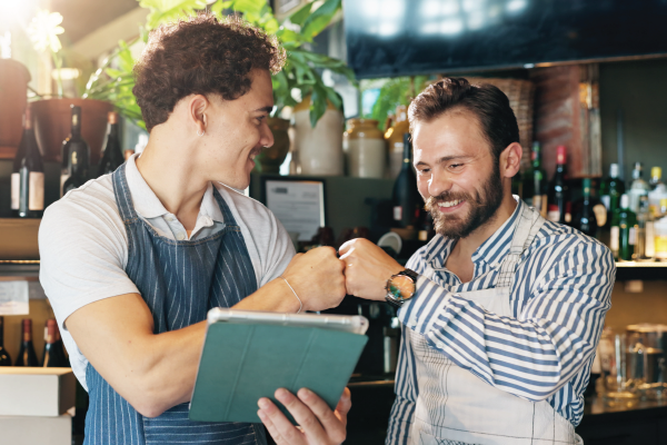 Two smiling baristas give each other a fist bump. One holds a tablet, both wear aprons inside a bar.