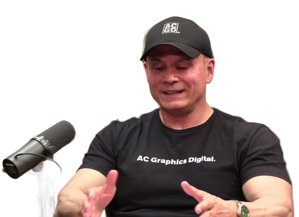 Man in black shirt and cap gesturing, seated near a microphone, white background.
