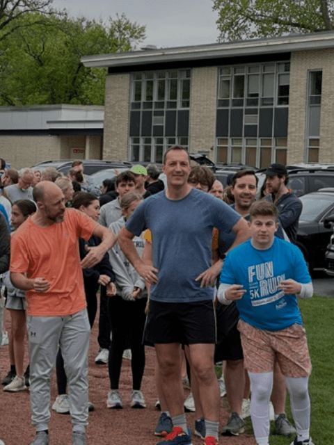 Group of people standing outside a brick building. Some wear athletic gear, arms out or hands on hips. Cars parked behind them.