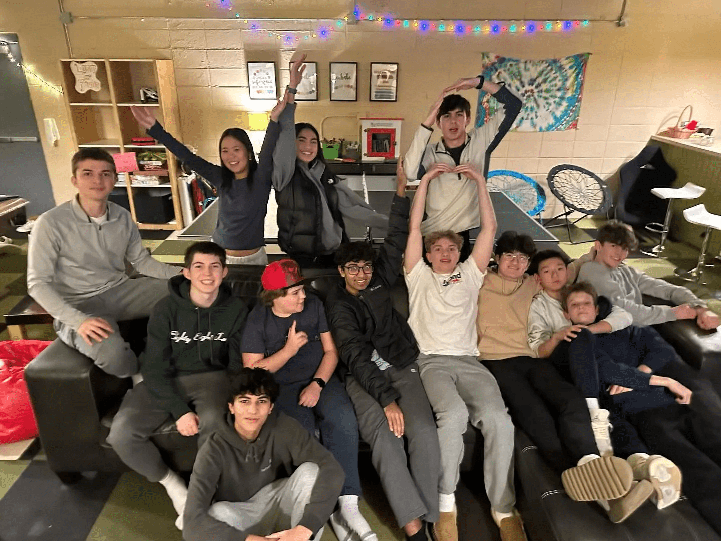 Group of teens posing on a long table, some waving and smiling, indoors with decorations.
