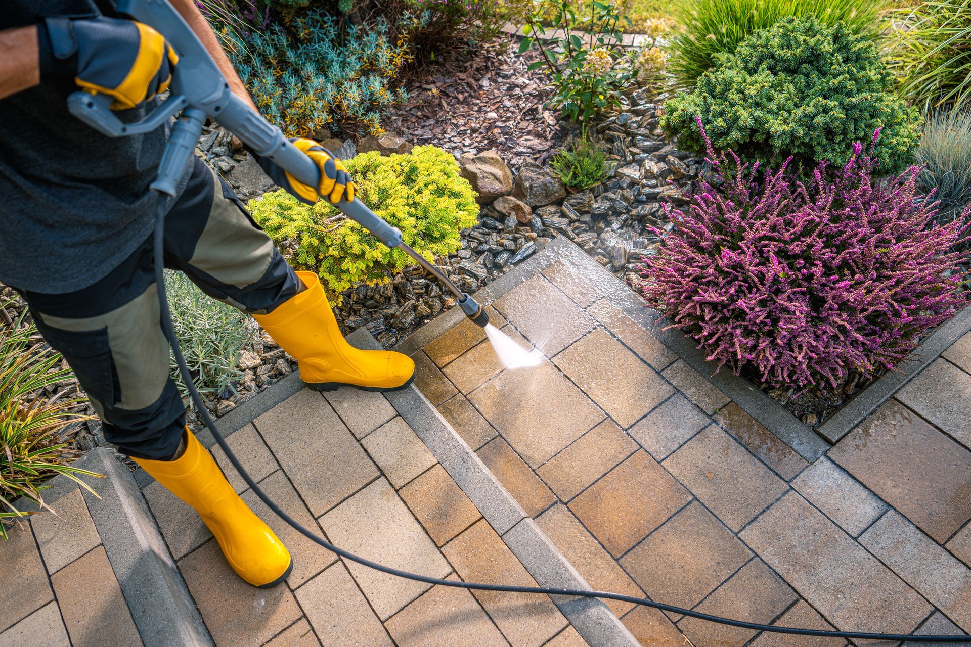 Person power washing stone steps in a garden, wearing yellow boots and gloves.