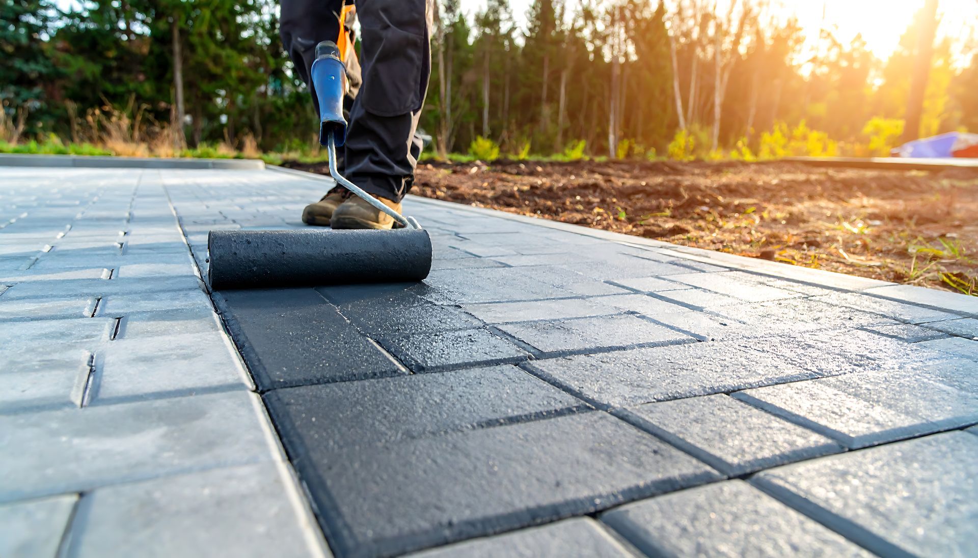 Worker applying sealant to brick pavers with a roller outdoors.