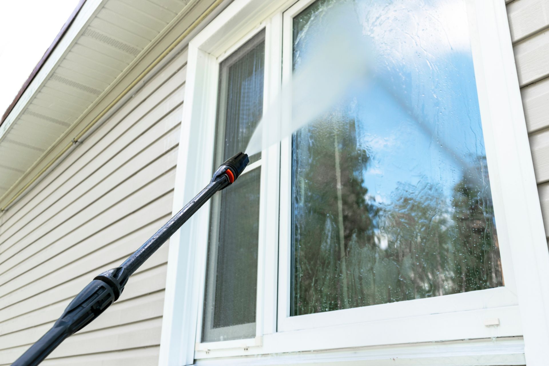 Pressure washer spraying water on a window, cleaning it, next to beige siding.
