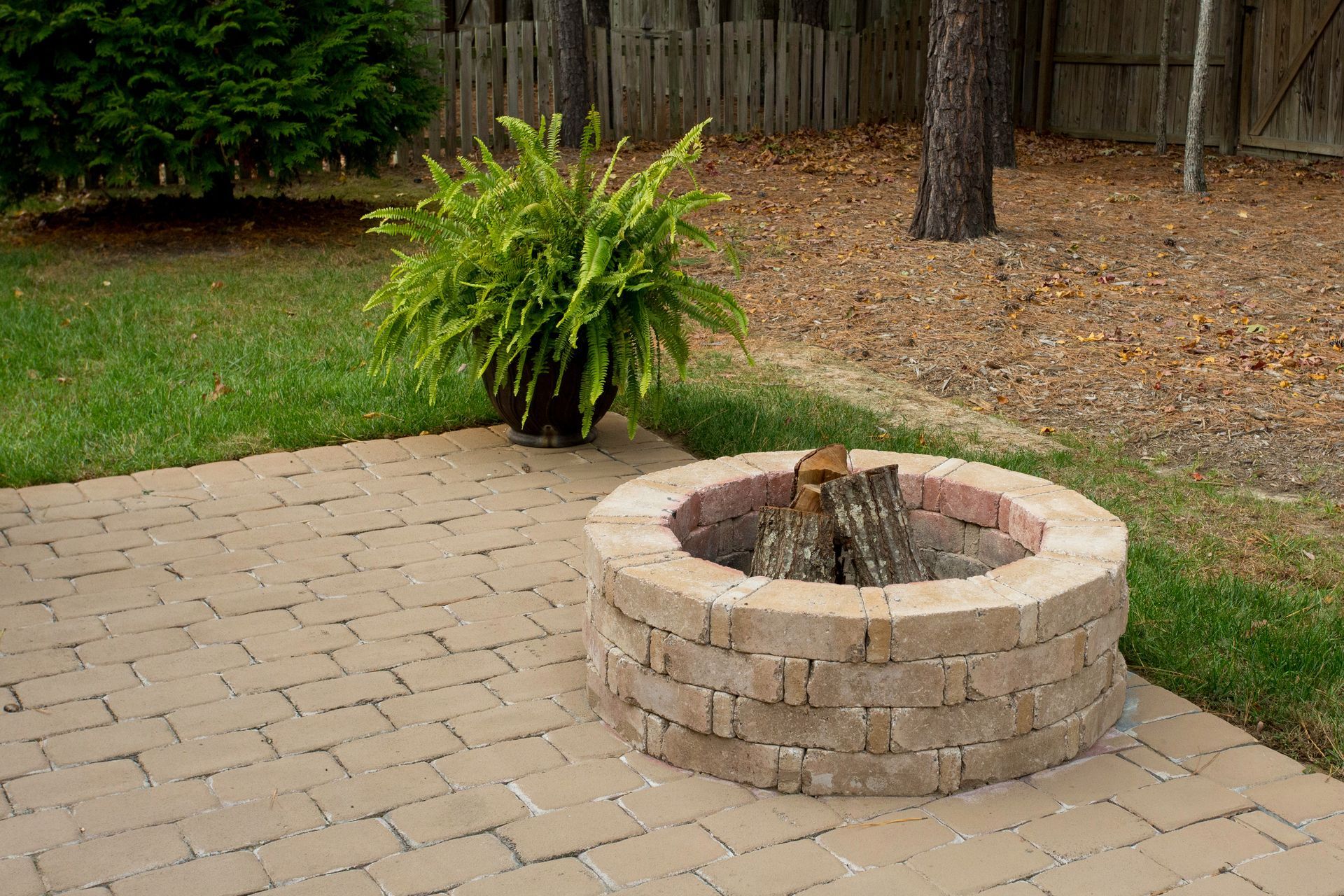 Brick fire pit with stacked logs on a brick patio with a fern and trees in the background.