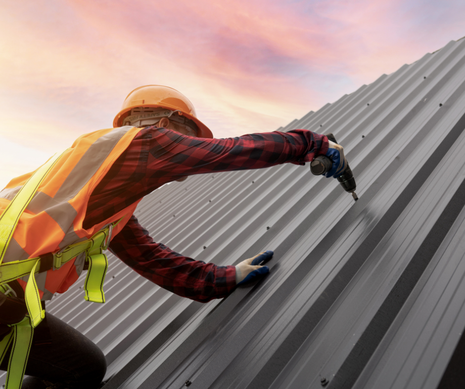 Roofer in safety gear using a drill on a metal roof, orange sunset in the background.