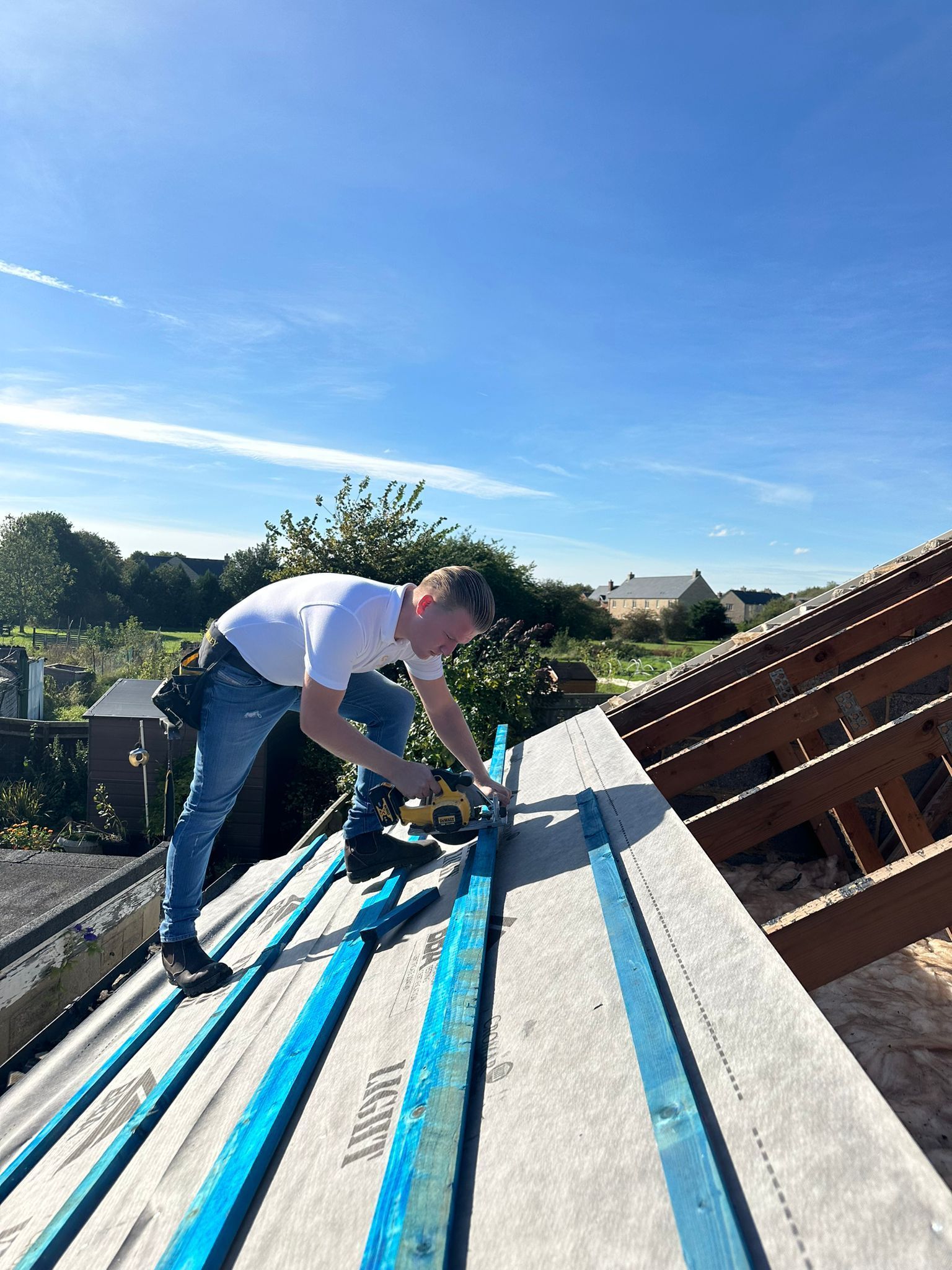 A person installs blue metal battens onto the gray, sloped roof of a house under a clear blue sky.