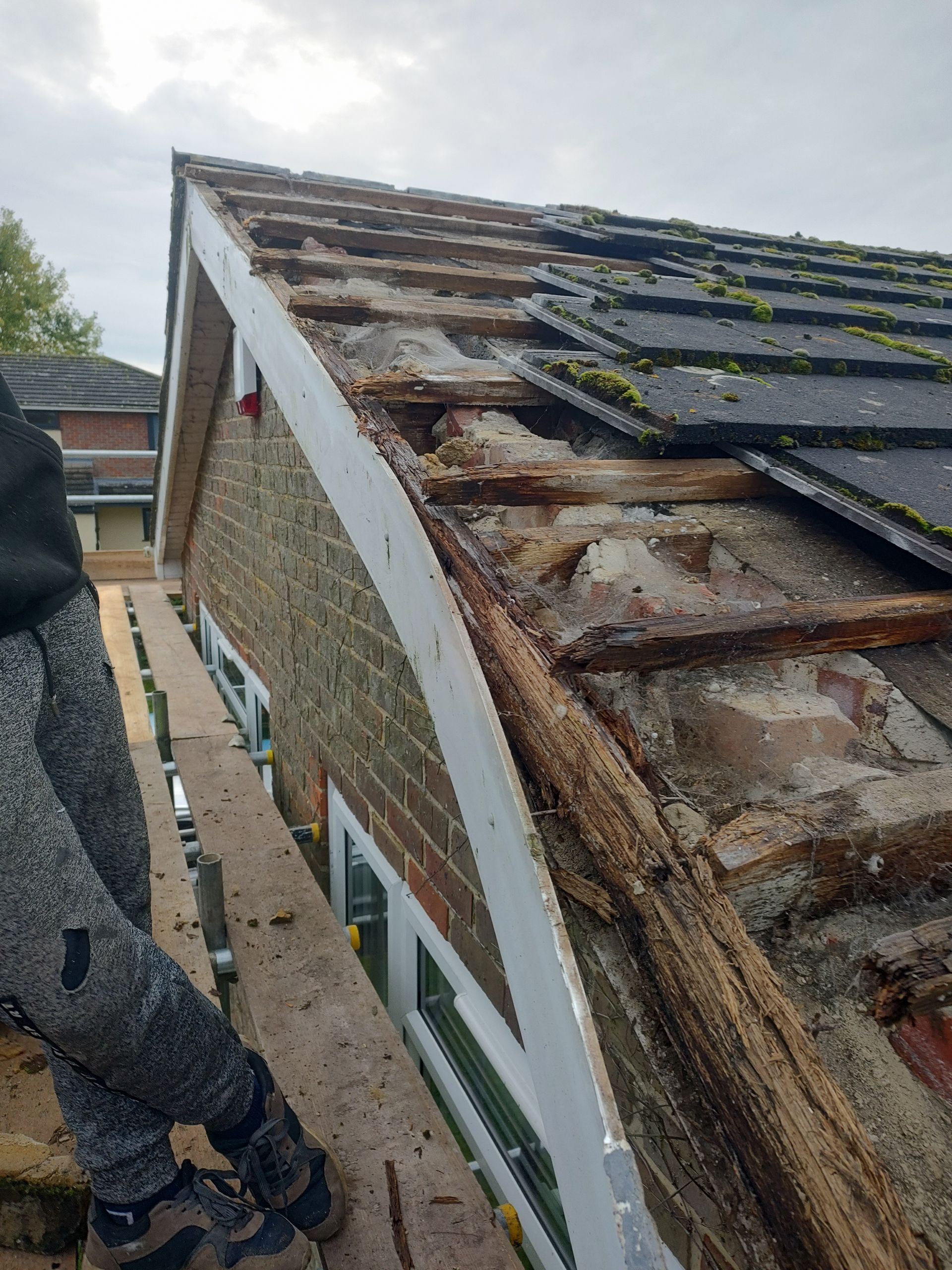 A person stands on scaffolding next to a residential roof undergoing repairs, showing damaged wooden rafters and tiling.