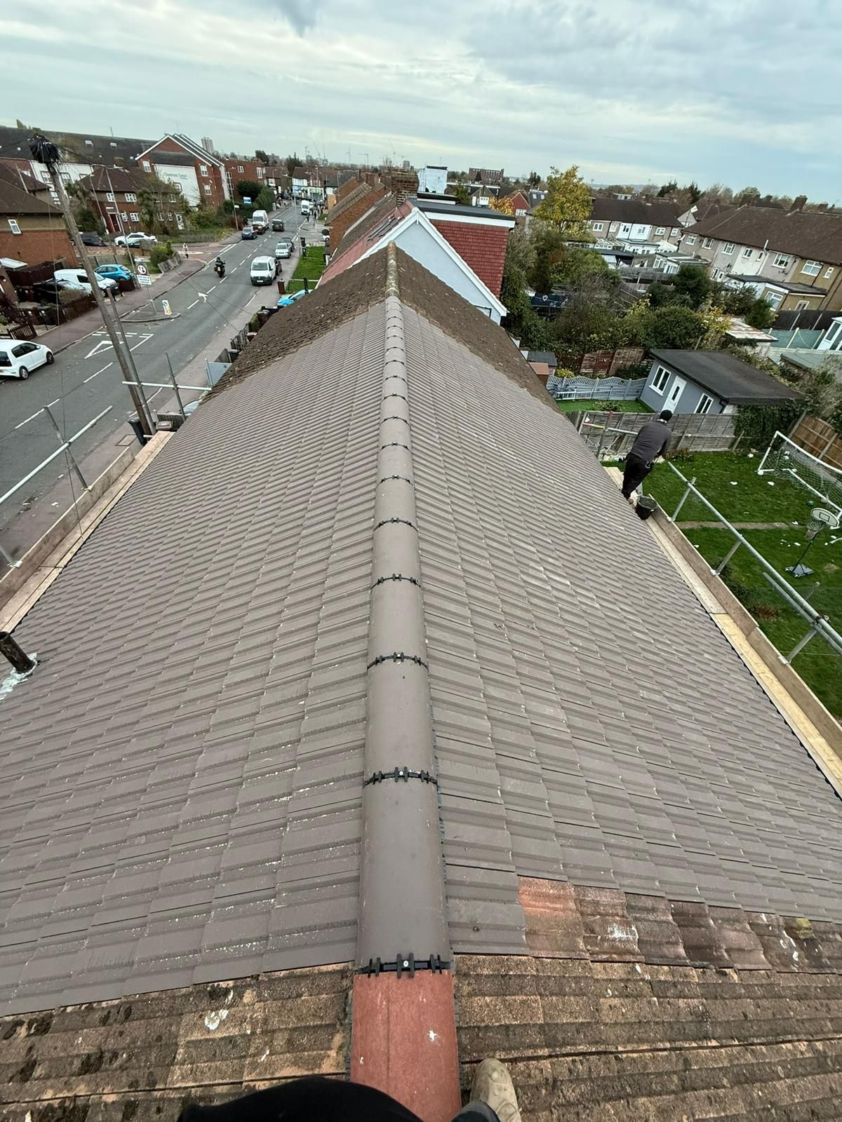 A view from a rooftop looking down the ridge of a house featuring a new grey tiled roof and a person working nearby.