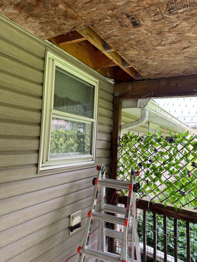Exterior view of a house with exposed ceiling, window, ladder, and greenery on a deck. Beige siding and trim.
