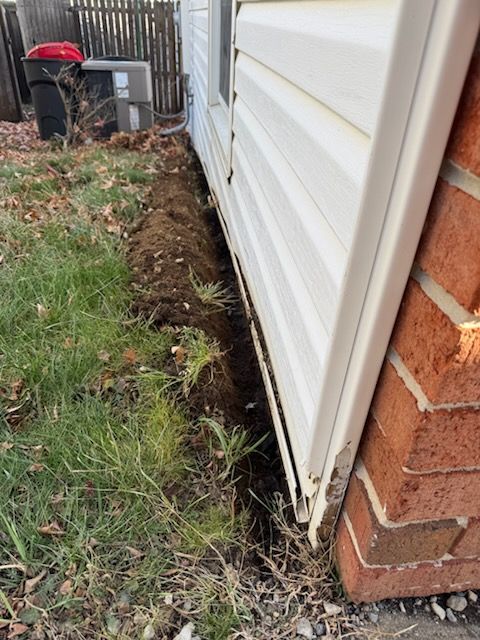 Trench dug next to a brick and siding exterior wall of a house, with grass in the foreground.