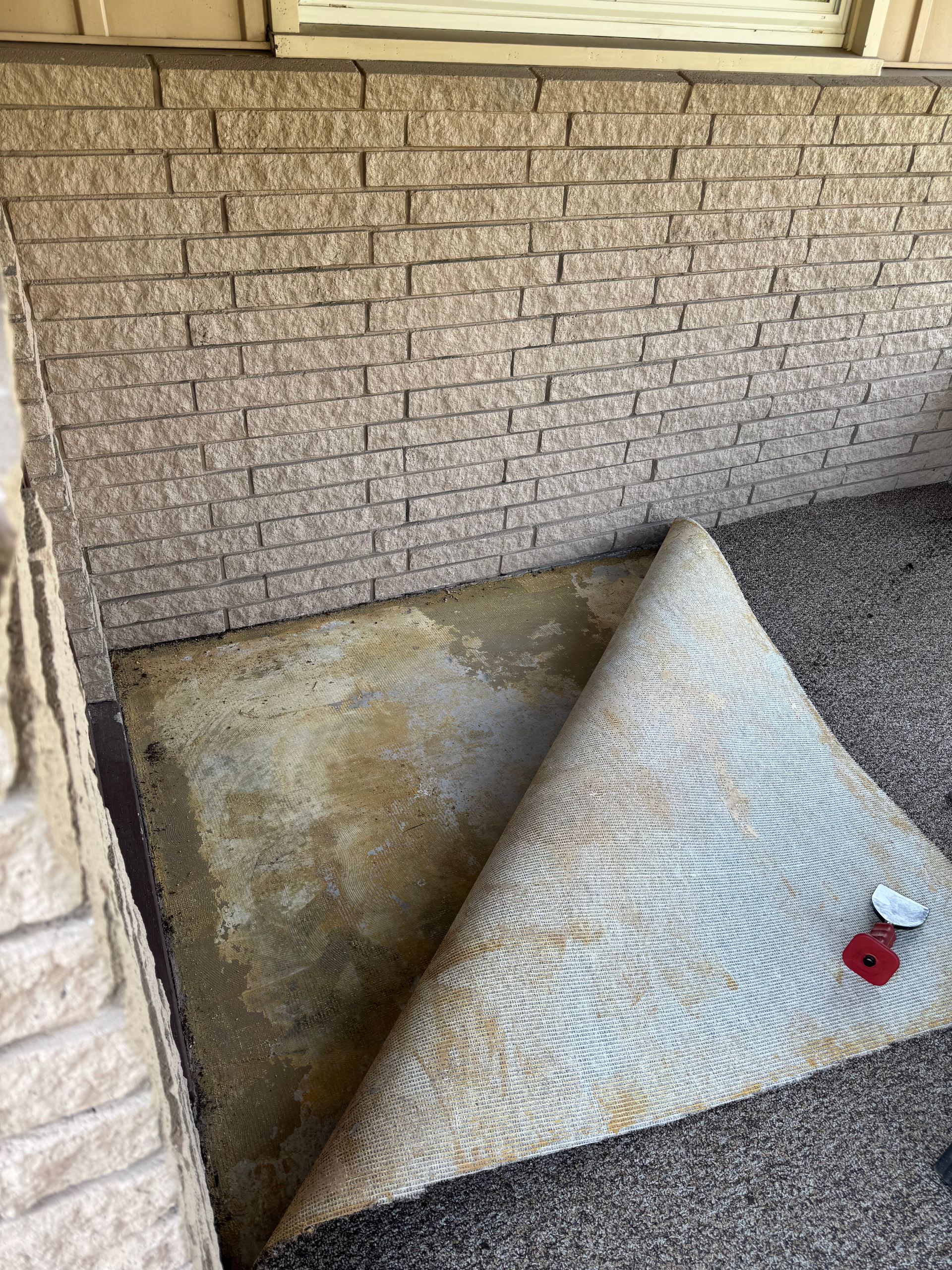 Beige brick wall and concrete floor. A rolled up patterned rug lies on the ground with a red object on it.