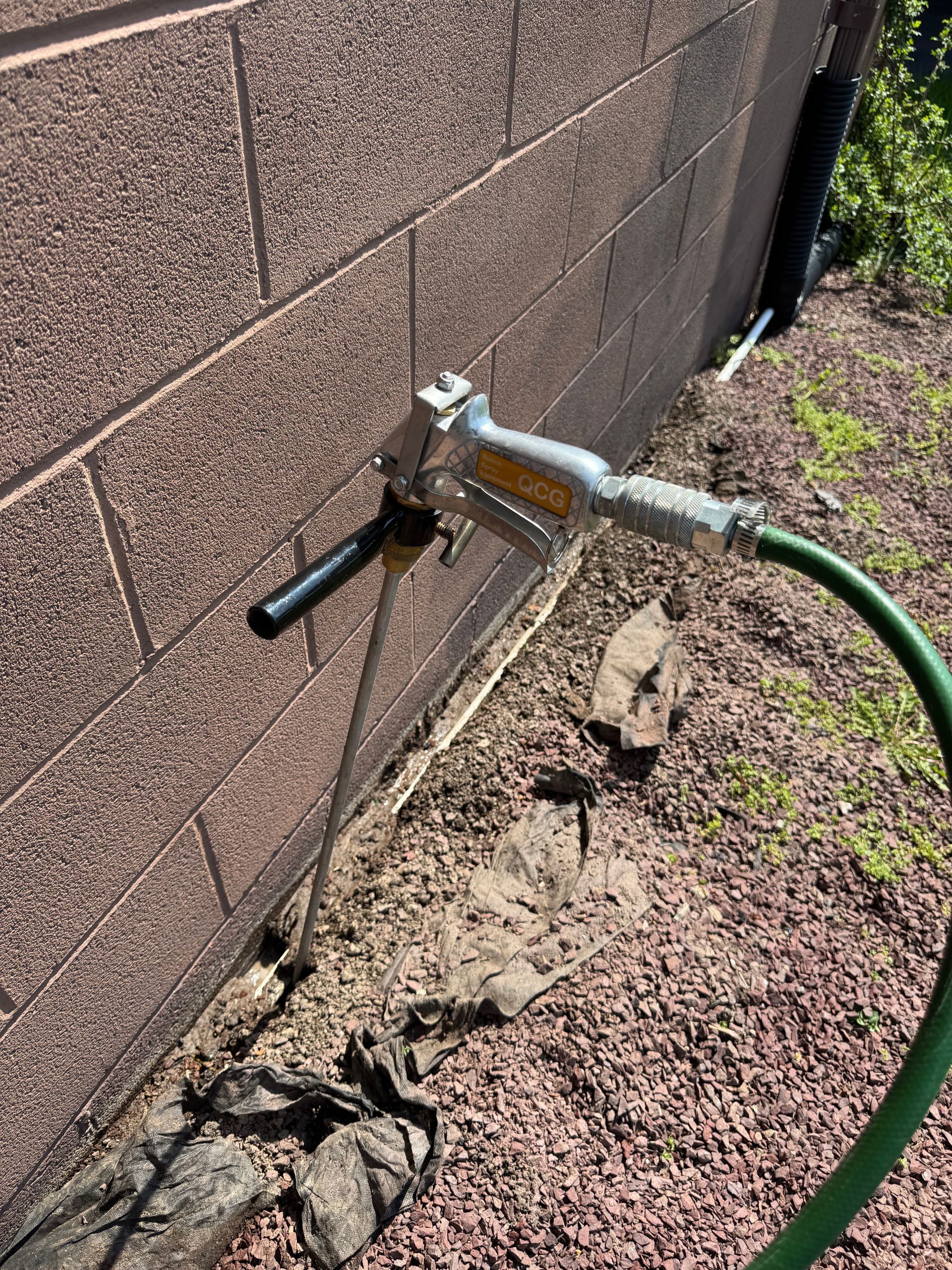 Sprayer attached to a hose pointed at a block wall. Ground covered in rocks and dirt.