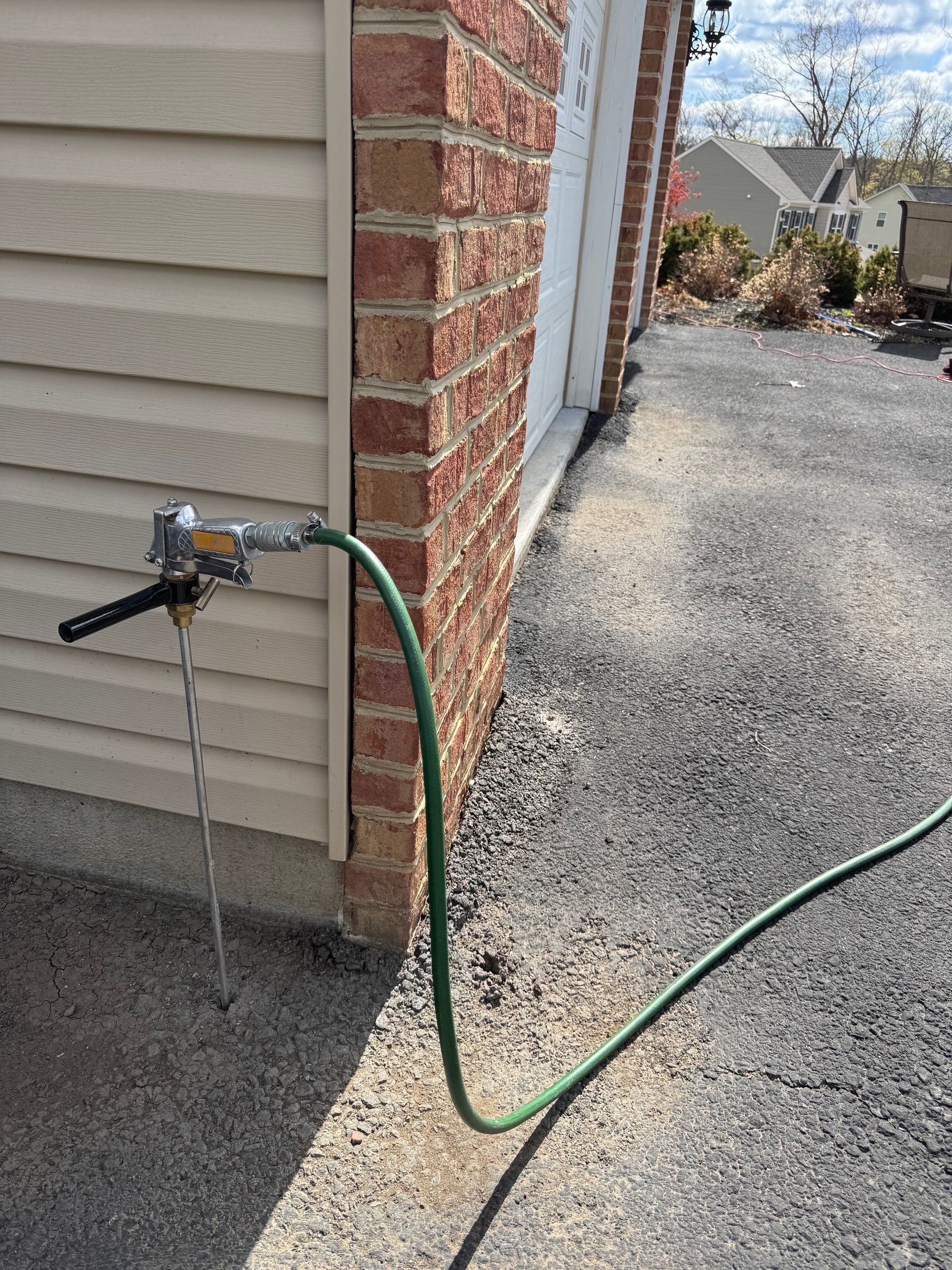Exterior water faucet and hose attached to a building with beige siding and a brick wall, on gravel.