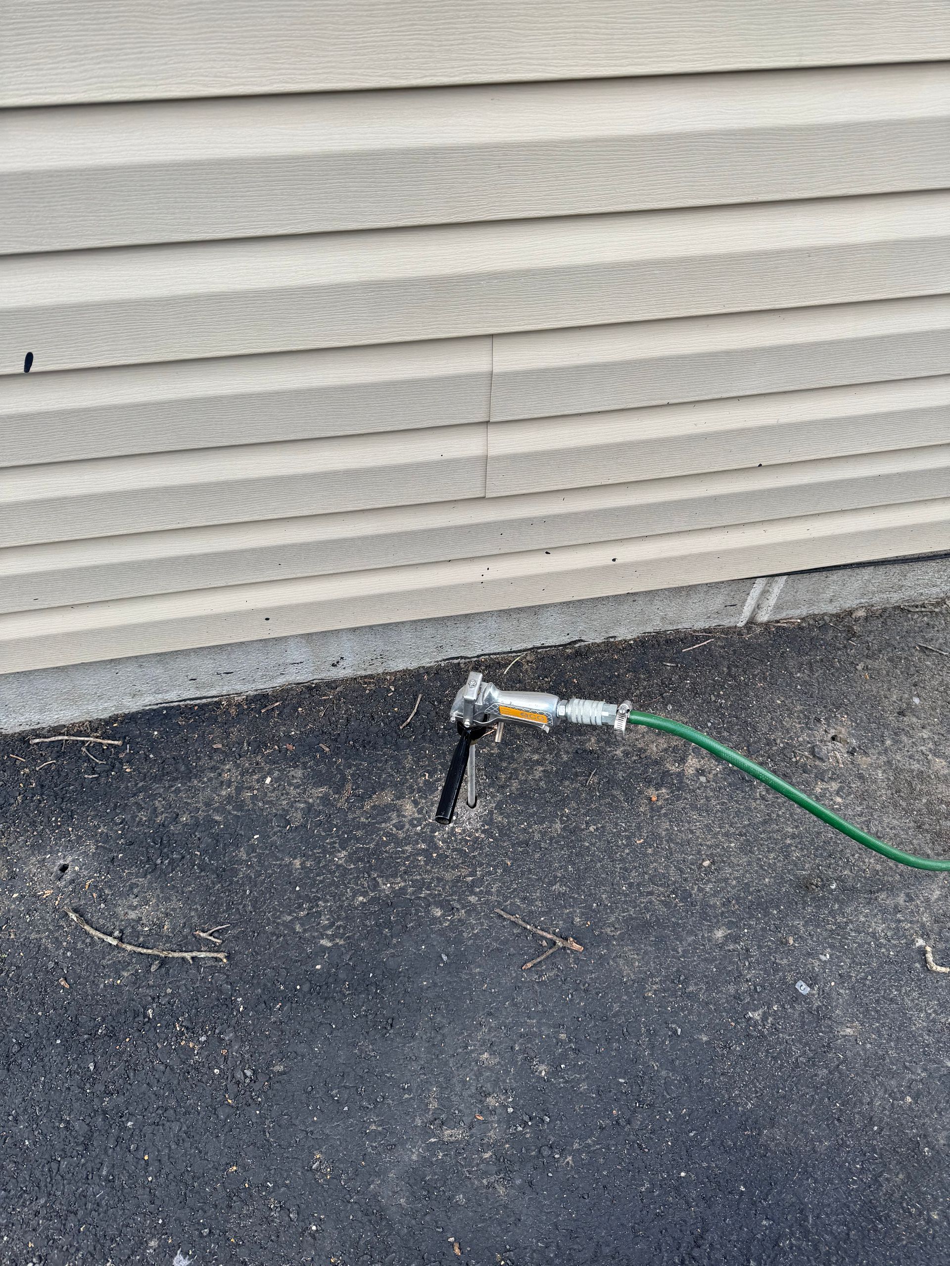 Green garden hose connected to a spigot on the side of a building, next to asphalt.