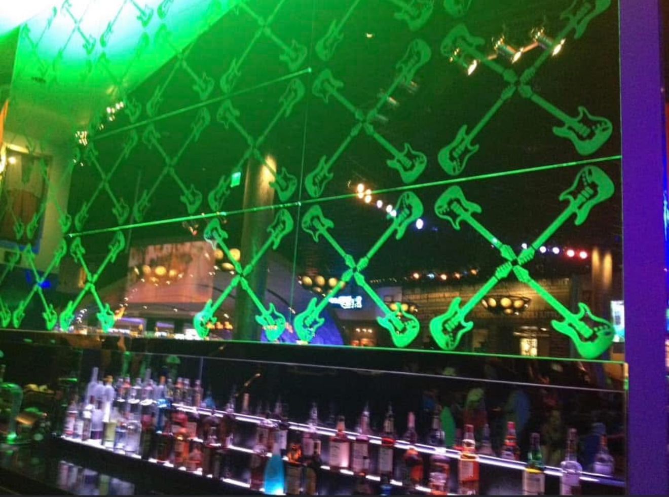 Bar with green-lit guitar silhouettes on the mirrored wall, bottles of liquor in front, and green ambient lighting.